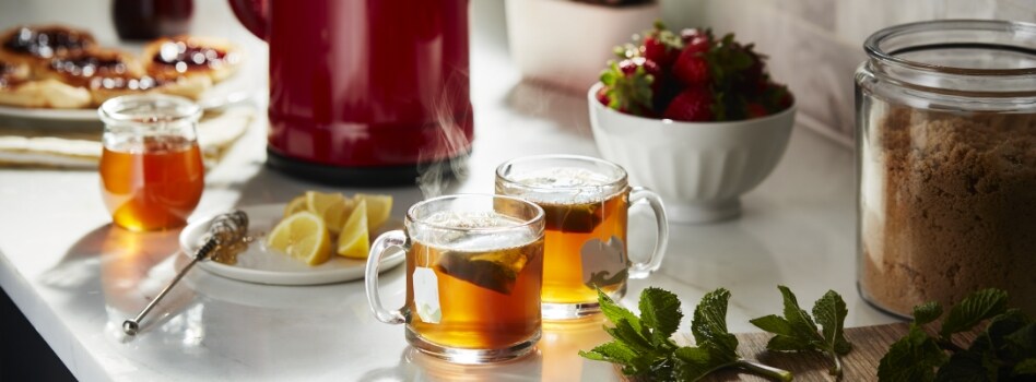 Items on a counter: a plate of tarts, a small container of honey, a red KitchenAid Kettle, a plate of lemon wedges, two cups of tea, a bowl of strawberries. another bowl, some mint leaves and a container of brown sugar. 